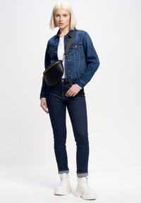 Jeansjakna v temno modri barvi z dvema sprednjima žepoma, bela majica, temne kavbojke in bele platforme. črn crossbody nahrbtnik izpostavlja oblikovanje.