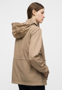 Γυναίκα που φοράει ανοιχτό καφέ μπουφάν με κουκούλα, με ελαστικά μανίκια και πλαϊνές τσέπες, φαίνεται από πίσω και στρέφει το κεφάλι της προς τα δεξιά.
