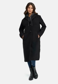 Long manteau noir en laine avec fermeture à boutons, deux poches avant et capuche. Porté par-dessus un sweat à capuche gris, associé à un jean bleu et des bottes noires.