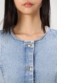  Lichtblauwe denimjacket met ronde hals, knoopsluiting aan de voorkant met zilveren knopen en zichtbare stikseldetails.