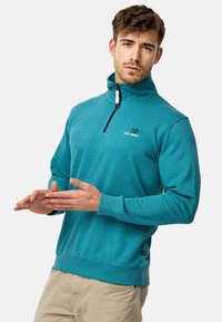 Sudadera de cremallera turquesa con cuello alto, logo bordado en el pecho y puños acanalados. El modelo posa con las manos abiertas, vistiendo pantalones claros.