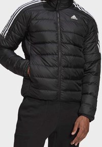 Homme portant une veste matelassée noire Adidas avec des détails blancs à trois bandes sur les manches et un pantalon noir, les mains partiellement dans les poches.