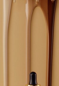 Trois flacons compte-gouttes contenant des liquides dans des nuances de beige et de marron, présentant des accents dorés et des pipettes noires, sur un fond beige.