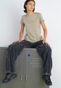 Šedé tričko s krátkym rukávom a logom, v kombinácii s voľnými čiernymi džínsami. Model sedí na sivých skrinkách a má na sebe čierne tenisky.