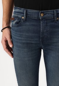 Sötétkék farmernadrág slim fit szabással,kopottas hatással, aranyszínű gombbal és bőrből készült folttal az elülső zseb közelében.