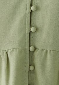 Lichtgroene, gestructureerde stof met een knoopsluiting aan de voorkant, voorzien van drie ronde stoffen knopen en een geplooid gedeelte aan de onderkant.