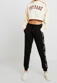 Krátká krémová mikina Ivy Park s červeným textem, doplněná černými tepláky s bílými logy po stranách a dokončená bílými teniskami.