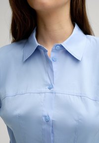 Hellblaues, kurzärmliges Blusenshirt mit spitzem Kragen, das eine Knopfverschlussleiste und subtile Stickdetails über der Brust aufweist.
