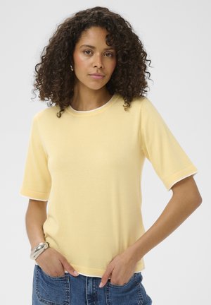 Femme aux cheveux bouclés, portant un haut jaune clair à manches courtes et un jean bleu, posant les mains dans les poches devant un fond uni.