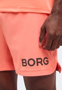 Κοντινό πλάνο ενός ατόμου που φοράει κοραλλί σορτς με το «BORG» τυπωμένο μαύρο στη αριστερή μηροκήλη, και ένα χέρι που ακουμπά δίπλα στα σορτς.