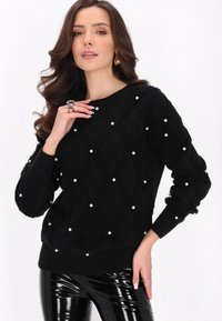 Schwarzer, flauschiger Pullover mit Perlenverzierungen, Rautenmuster, rundem Halsauschnitt und gerippten Bündchen. Getragen mit glänzenden schwarzen Hosen.