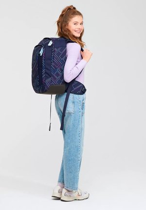 Navyblauer Rucksack mit geometrischen Musterelementen, gepolsterten Trägern und einem schwarzen Boden. Modell trägt ein helllila Oberteil und hellblaue Jeans.