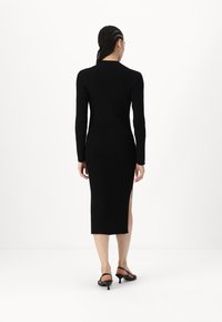 Abito nero a costine con maniche lunghe, lunghezza midi, spacco laterale, silhouette aderente, indossato con sandali neri con cinturini, design minimalista.