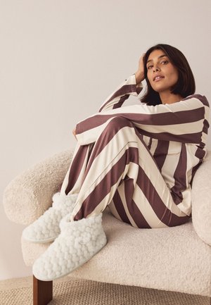 Femme assise sur un fauteuil blanc texturé, portant un pyjama rayé marron et blanc et des chaussons bottines blancs et duveteux, l'air détendue et légèrement souriante.