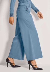 Vrouw in een wijde blauwe broek en zwarte hoge Mary Jane schoenen die tegen een effen lichte achtergrond loopt.