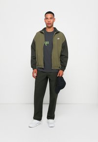 Olivnozeleni vetrovka s črnimi detajli, siva majica s_logotipom, usklajene olivno zelene cargo hlače in bele superge. Temna kapa v roki.