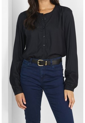 Femme portant un chemisier noir à manches longues boutonné, rentré dans un jean bleu foncé taille haute avec une ceinture noire ornée d'une boucle dorée.