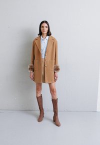 Cappotto di lana color cammello con bottoni grandi e dettagli in pelliccia sulle maniche, abbinato a una camicia blu, una gonna beige e stivali al ginocchio marroni.