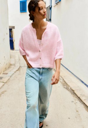 Femme marchant dans une rue étroite portant un cardigan rose boutonné, un jean large bleu clair et des sandales noires.