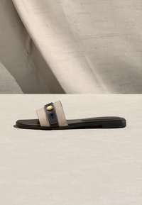 Επίπεδη σανδάλι με πλατιά μπεζ υφασμάτινη λουρίδα που έχει μαύρη επένδυση και χρυσό κουμπί, τοποθετημένη σε ουδέτερη επιφάνεια.