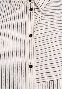 Chemise à boutons avec un fond blanc et de fines rayures verticales noires, dotée de boutons noirs et d'une poche plaquée sur le côté gauche.