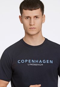 Donkerblauwe T-shirt van katoen met de tekst "COPENHAGEN LINDBERGH" in lichtblauw en wit. Korte mouwen, ronde hals.