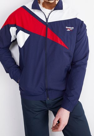 Mand iført navyblå, rød og hvid Reebok lynlåstrøje med geometrisk design, kombineret med mørke bukser, stående mod en hvid baggrund.