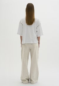 Weißes oversized T-Shirt mit dekorativen Ösen an den Schultern, kombiniert mit hellbeigen weiten Hosen mit strukturierten vertikalen Streifen.