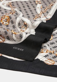 Pañuelo de seda con un patrón de logotipo en beige y negro, cadenas doradas, un colgante en forma de corazón y un borde negro con el texto "GUESS" en blanco.