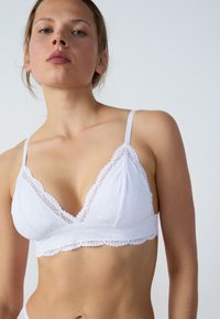 Bralette en dentelle blanche avec un design de bonnet triangulaire, des bretelles ajustables, des bords festonnés et des motifs texturés sur le tissu.