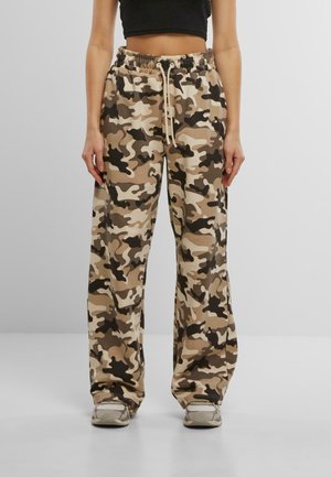 Kvinde iført løstsiddende camouflagebukser i beige, brunt og sort med snøre i taljen og grå sneakers, stående mod en ensfarvet baggrund.