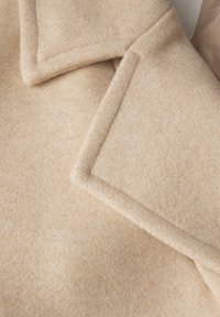 Tessuto misto lana beige con una texture morbida, caratterizzato da un ampio colletto e bordi strutturati, che enfatizza linee pulite e un design sartoriale.