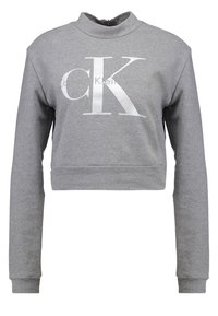 Šedá zkrácená mikina s dlouhými rukávy a vysokým límcem. Obsahuje velké bílé tištěné logo "CK" a strukturovanou tkaninu.