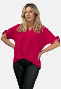 Fuksjowa koszulka z krótkim rękawem i dekoltem w serek, z podwiniętymi mankietami i z przodu z detalem w formie skrętu. W połączeniu z dopasowanymi czarnymi spodniami.