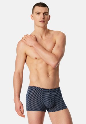 Gespiereerde man met kort haar, gekleed in marineblauwe en wit gestreepte boxershorts, staand tegen een effen lichte achtergrond.