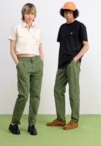 Two individuals pose together; one wears a cream cropped shirt and olive pants, the other a black t-shirt and olive pants. Both have casual shoes.
