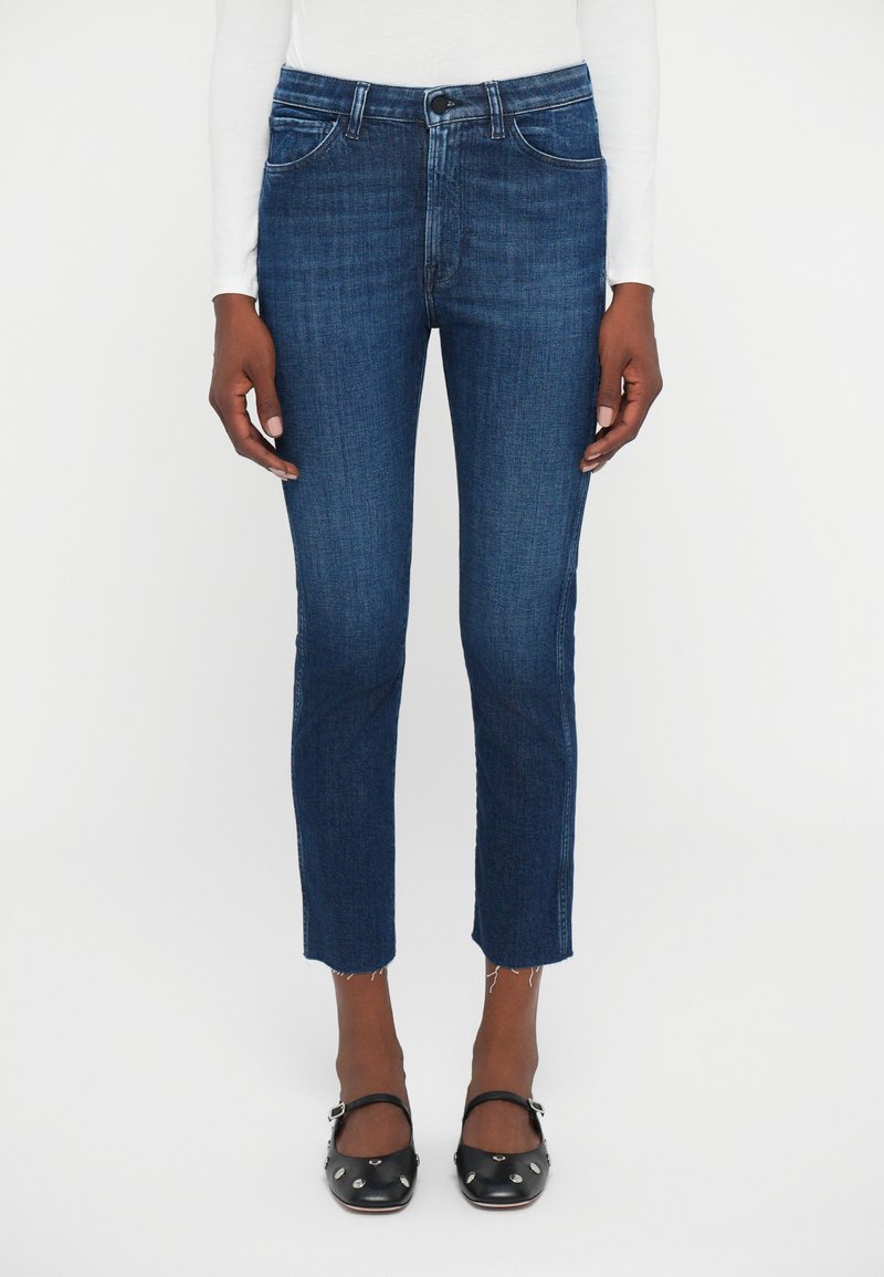 Jeans skinny blu scuro cropped con orlo grezzo indossati da una persona con scarpe Mary Jane nere con borchie e maglietta bianca a maniche lunghe.
