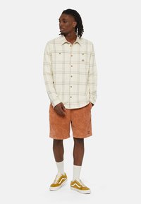 Camisa de cuadros en crema con líneas azules y amarillas, combinada con pantalones cortos naranjas. Las características notables incluyen botones en la camisa y un logo de acento.