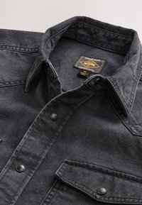 Black denim jacket with pointed collar, snap buttons, chest pocket with flap, and a branded label inside the collar.