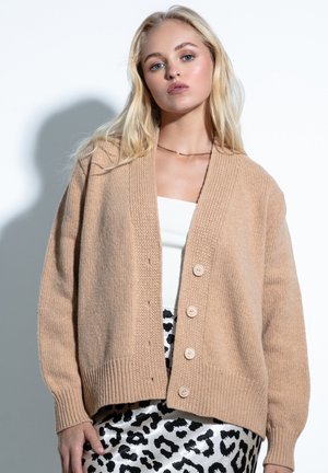 Beżowy sweter z dzianiny z prążkowanymi mankietami i dołem, z pięcioma guzikami z przodu, noszony na białą bluzkę i spódnicę w czarno-białą panterkę.