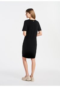 Czarna sukienka z krótkim rękawem i okrągłym dekoltem, gładka w dotyku, z małym okrągłym akcentem z tyłu, zestawiona z beżowymi butami na platformie.