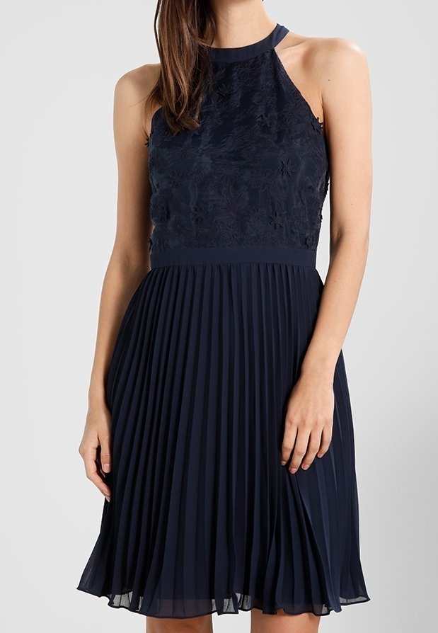 Navy blue dress with a fitted floral lace bodice and textured pleated skirt. Straps form a halter neckline; knee-length flowy design.