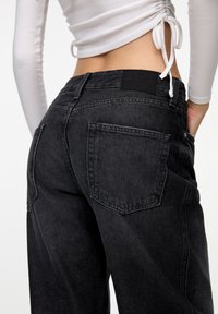 Jean en denim noir à coupe décontractée, avec deux poches arrière, une étiquette à la taille et un tissu texturé. Le design est simple et classique.