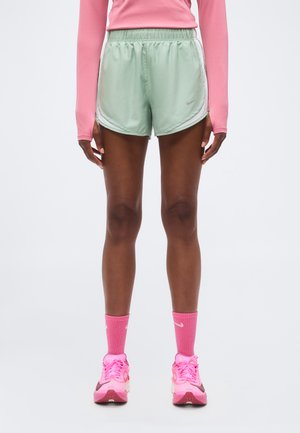Modell trägt hellgrüne Sportshorts, pinkes Langarmshirt, pinke Socken und pinke Laufschuhe und steht vor einem weißen Hintergrund.