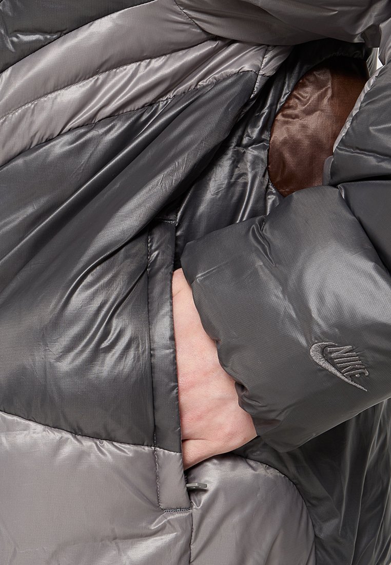 Chaqueta acolchada gris con un diseño acolchado, que presenta paneles brillantes y mates, y un detalle de bolsillo. Incluye un pequeño logo de Nike en la manga.