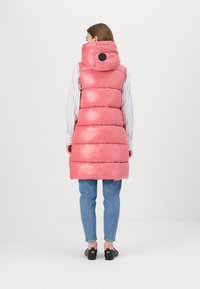 Gilet puffoso rosa con cappuccio, caratterizzato da un'imbottitura orizzontale, un accento circolare nero sul retro e jeans in denim blu a contrasto.