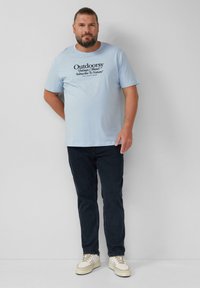 Lichtblauwe katoenen T-shirt met zwarte tekstgraphics die "Outdoorsy" en "Abonneer je op de Natuur" zeggen. Gecombineerd met donkerblauwe jeans en witte sneakers.