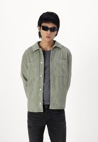 Chaqueta de pana verde con dos bolsillos en el pecho y un cuello, abotonada sobre una camisa gris oscuro, combinada con jeans negros desgastados y gafas de sol.