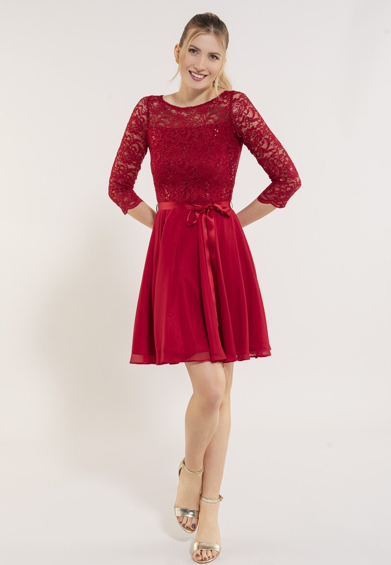 Robe rouge avec un corsage en dentelle à manches trois-quarts, une ceinture en satin, et une jupe en chiffon fluide. Talons avec bride de cheville dorés.