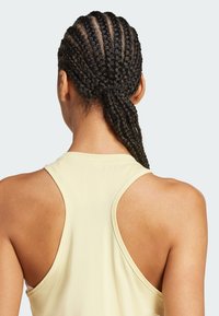 Top de deporte color amarillo claro con diseño de espaldas al descubierto, que presenta una tela texturizada y una espalda baja que muestra el cabello trenzado.